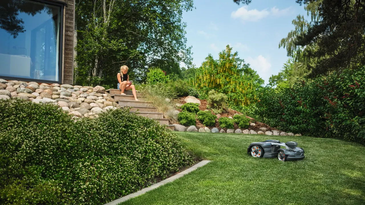 Robot tallagespa Husqvarna Automower® tallant la gespa d'un jardí amb parterres i escales de pedra